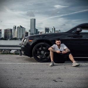 Фото мужчины с татуировками на руке, сидящего рядом с черным Rolls-Royce. Городской пейзаж на заднем плане.