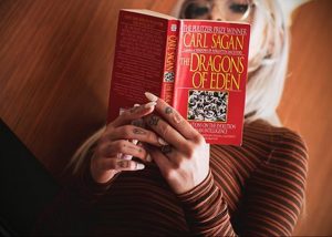 Девушка с длинными светлыми волосами держит книгу перед собой, демонстрируя татуировки на руках.