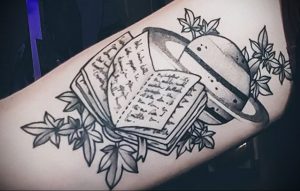 Фото тату на руке: открытая книга и планета Сатурн, черно-белая татуировка, детализированный рисунок. Оригинальный и символичный образ.