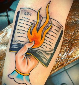 Фото тату на руке: горящая книга с цифрой '451' на обложке, рука держит книгу. Яркие цвета, стиль олдскул.