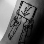 Фото тату на руке с изображением рук, держащих открытую книгу, из которой растут цветы. Минималистичный стиль, тонкие линии и нежный дизайн.