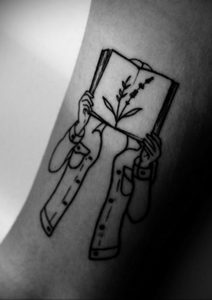 Фото тату на руке с изображением рук, держащих открытую книгу, из которой растут цветы. Минималистичный стиль, тонкие линии и нежный дизайн.