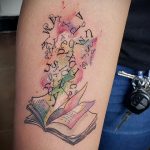 Фото тату на руке с изображением раскрытой книги, из которой вылетают буквы. Акварельный стиль, яркие цвета и динамичная композиция.