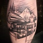 Фото тату на ноге с изображением открытой книги, из которой вытекает водопад, а на фоне видны горы и лес. Выполнено в черно-серых тонах.