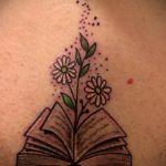 Фото тату на спине с изображением открытой книги, из которой растут ромашки, и летящие вокруг них точки. Выполнено в ярких цветах.