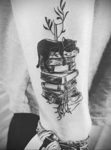 Фото тату на ноге с изображением спящего черного кота, лежащего на стопке книг, с веткой дерева над ним. Выполнено в графическом стиле.