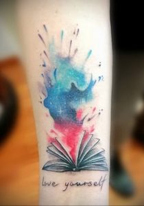 Фото тату на руке: раскрытая книга, из которой вырывается звездное небо, выполнена в акварельной технике. Красивый рисунок татуировки для мечтателей и любителей космоса.