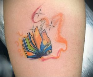 Фото тату на руке: раскрытая книга, излучающая свет и искры, выполнена в ярких оранжевых и желтых тонах. Оригинальный рисунок татуировки для ценителей литературы.