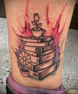 Фото тату на предплечье: стопка книг с чашкой кофе и цветком, выполненная в ярких цветах с брызгами краски. Идеальный эскиз тату для любителей чтения.