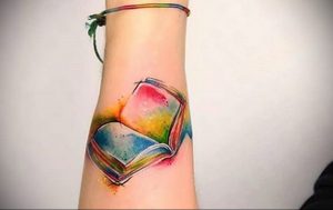 Фото тату на предплечье: открытая книга с яркими, радужными страницами, выполненная в акварельной технике. Нежный и красочный рисунок татуировки.
