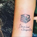 Фото тату на предплечье: небольшая стопка книг в серо-черных тонах с цитатой 'I have lived a thousand lives'. Изящный рисунок татуировки.