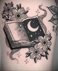 Фото татуировки в стиле blackwork, изображающей открытую книгу с луной внутри, окруженную цветами и звездами.