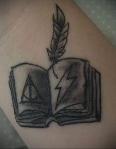 Фото татуировки в стиле blackwork, изображающей открытую книгу с символами Гарри Поттера, пером и молнией.