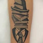 Фото тату на руке: стопка книг, на которой стоит шляпа, а под ней – силуэт мужчины в костюме и галстуке. Черно-белый рисунок татуировки.