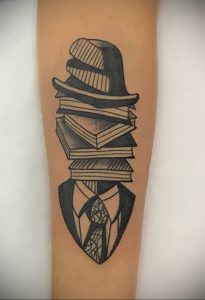Фото тату на руке: стопка книг, на которой стоит шляпа, а под ней – силуэт мужчины в костюме и галстуке. Черно-белый рисунок татуировки.