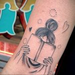 Фото минималистичной татуировки на руке с изображением девушки, читающей книгу. Вокруг девушки изображены точки и сердечки.