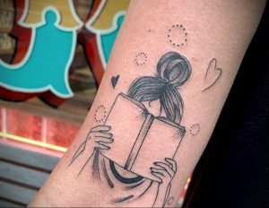 Фото минималистичной татуировки на руке с изображением девушки, читающей книгу. Вокруг девушки изображены точки и сердечки.