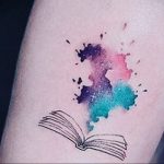 Фотография татуировки на руке с изображением раскрытой книги, окруженной красочными акварельными брызгами.