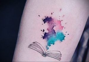 Фотография татуировки на руке с изображением раскрытой книги, окруженной красочными акварельными брызгами.