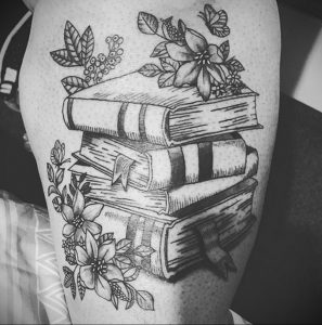 Фотография татуировки на бедре с изображением стопки книг, украшенной цветами и листьями.