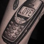 Черно-белое фото тату на предплечье с изображением старого телефона, на экране которого написано слово LOVE. Стиль татуировки - реализм.