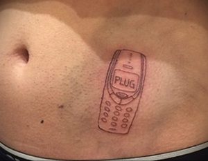 Фото тату на животе: старый кнопочный телефон с надписью 'PLUG' на экране. Простой и минималистичный рисунок в черных тонах.