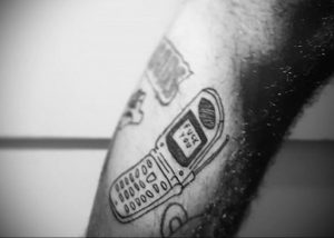 Фото тату на руке: старый кнопочный телефон с надписью 'FUCK YOU' на экране. Рисунок выполнен в стиле doodle, с использованием тонких линий.