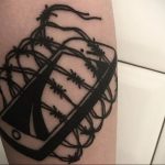 Черно-белая татуировка на ноге с изображением смартфона, обмотанного колючей проволокой. Символическая татуировка, отражающая зависимость и ограничение свободы.