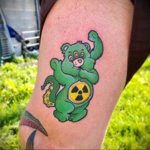 Фото тату на ноге: зеленый мишка Care Bear с символом радиации на животе, выполненный в ярком и мультяшном стиле.