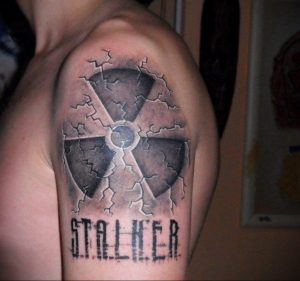 Фото тату: знак радиации с трещинами и надписью "STALKER", выполненный в черно-серых тонах. Татуировка вдохновлена популярной видеоигрой.