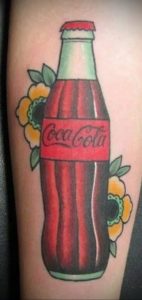 Фото татуировки на руке с изображением бутылки Coca-Cola, украшенной цветами. Рисунок выполнен в стиле олдскул, с использованием ярких цветов и четких контуров.