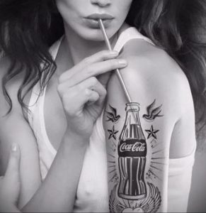 Фото татуировки на плече с изображением бутылки Coca-Cola, украшенной летящими птицами и звездами. Рисунок выполнен в черно-белой гамме, с использованием тонких линий и детализированной проработкой.