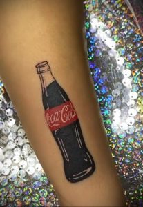 Фото тату бутылки Coca-Cola на руке. Черный контур, красная этикетка, реалистичный рисунок. Эскиз тату в стиле реализм.