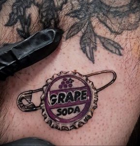 Фото тату: реалистичная татуировка крышки от Grape Soda, скрепленной булавкой. Цветная татуировка на теле.
