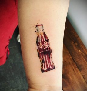 Фото тату: реалистичная акварельная татуировка бутылки Coca-Cola на предплечье, выполненная в красных и коричневых тонах. Идеально для любителей классического напитка.