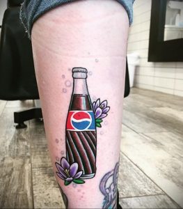 Фото тату на ноге с изображением бутылки 'Pepsi', украшенной цветами. Элегантный и стильный рисунок в ярких цветах.