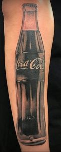 Фото тату на руке с изображением классической бутылки 'Coca-Cola'. Черно-белый рисунок с реалистичной прорисовкой.