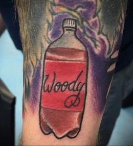 Фото тату на руке с изображением бутылки напитка 'Woody'. Красочный рисунок с детальной прорисовкой этикетки и формы бутылки.