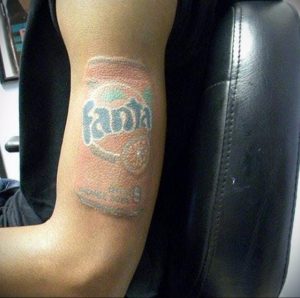 Фото тату на бицепсе с изображением старой банки Fanta. Рисунок выполнен в приглушенных тонах, с эффектом выцветшей краски, создавая винтажный вид.