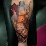 Фото тату на предплечье с изображением стеклянной бутылки газировки Castle Beverage, окруженной яркими осенними листьями и брызгами напитка.