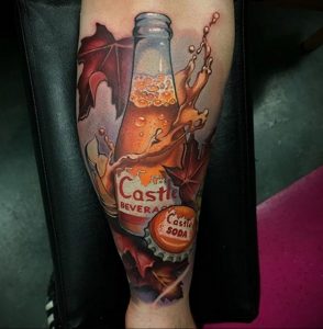 Фото тату на предплечье с изображением стеклянной бутылки газировки Castle Beverage, окруженной яркими осенними листьями и брызгами напитка.