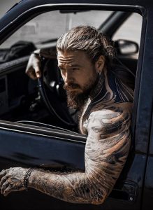Фотография мужчины с татуировками на руках, сидящего за рулем автомобиля. Рукава покрыты сложными узорами в черных тонах.