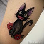 Фото тату на руке с изображением черного кота Дзиро из аниме 'Ведьмина служба доставки' в окружении красных цветов.