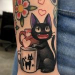 Фото тату на предплечье: черный кот держит чашку с котенком, окруженный цветами и сердечками в стиле old school.