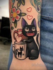 Фото тату на предплечье: черный кот держит чашку с котенком, окруженный цветами и сердечками в стиле old school.