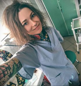 Фото тату на руках и шее у девушки-медсестры в голубом медицинском костюме. Разноцветные татуировки в стиле олдскул и традишн.