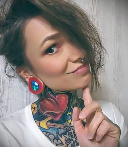 Фото медсестры с татуировкой на шее в виде сердца, пронзенного стрелой, и яркими сережками.