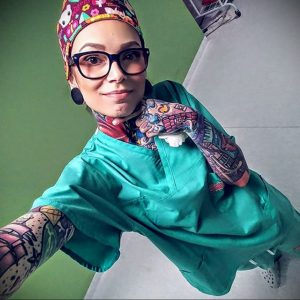 Фотография девушки-медика в медицинской форме и головном уборе с множеством татуировок на руках и шее. Яркие рисунки, демонстрирующие индивидуальность.