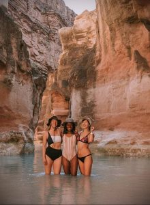 Фотография трех девушек в купальниках, стоящих в воде в каньоне, с татуировками на телах.
