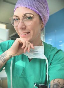 Фотография медицинской работницы в шапочке и маске, с задумчивым взглядом и татуировками на руках.